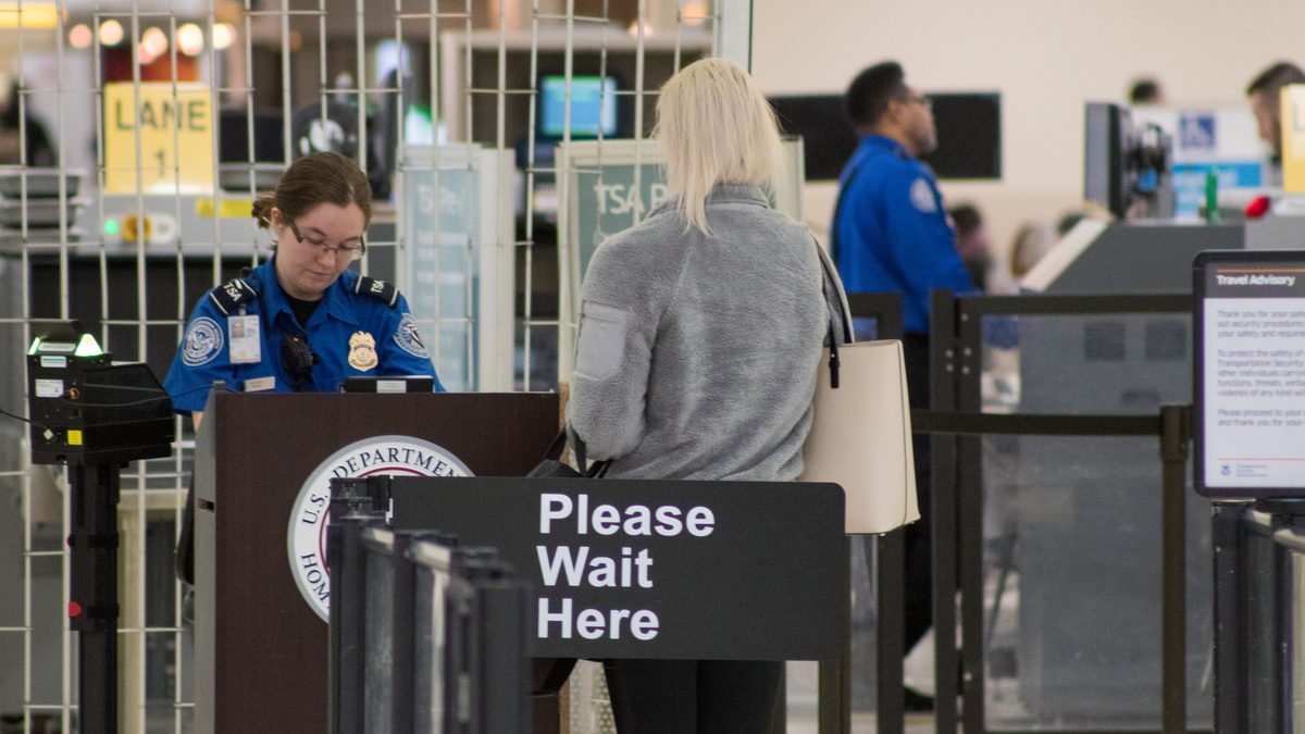A passenger waiting at a Please Wait Here sign while a TSA officer checks documents at an airport security checkpoint