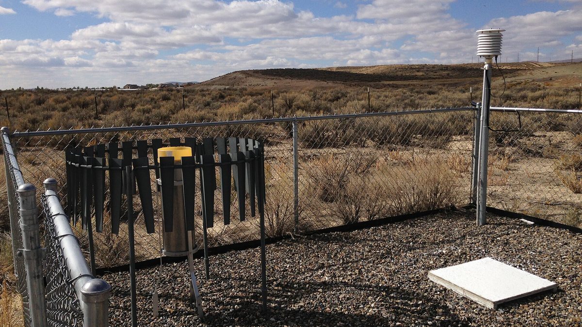 A fenced National Weather Service Cooperative Observer weather station with an alter-shielded precipitation gauge, a louvred thermometer shelter and a white snow board on gravel