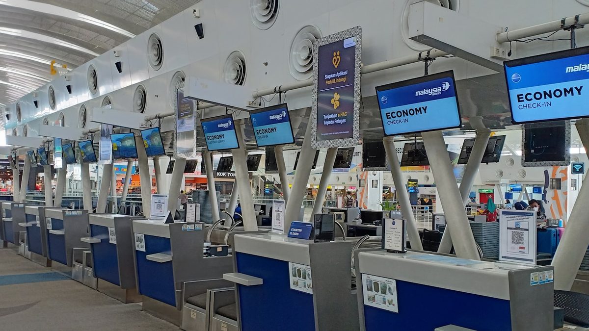 A row of airline check-in counters at an airport terminal, screens showing economy check-in