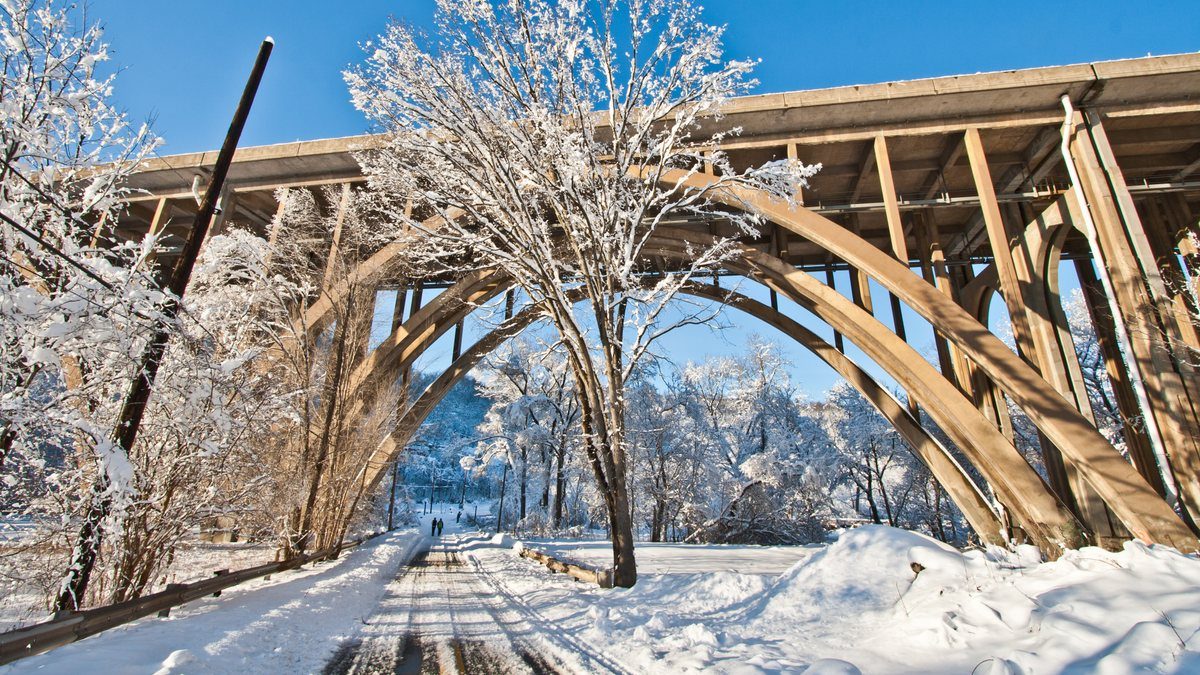 A snow-covered road running beneath the concrete arches of the Commercial Street Bridge in Pittsburgh on a clear winter day