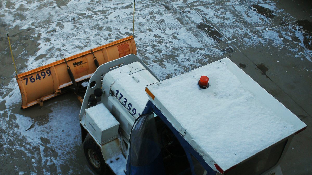 An orange snow plough blade attached to a small airline ground vehicle clearing a snow-covered aircraft apron at Pittsburgh International Airport