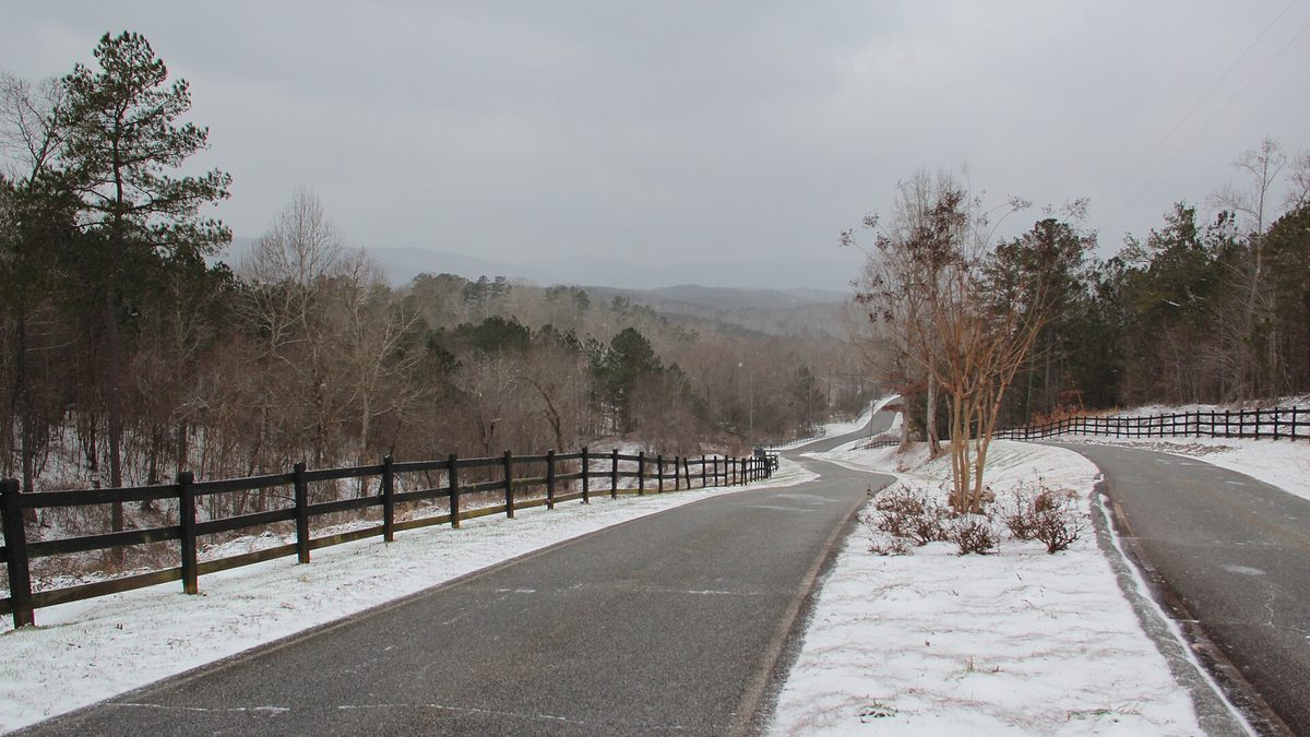 A rural road curving through north Georgia foothills with a thin dusting of snow on the grass verges and bare trees under a grey sky