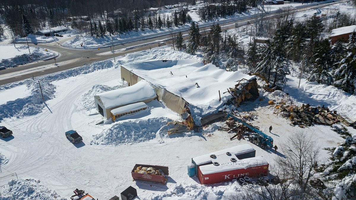 Aerial view of a school bus garage in Negaunee, Michigan with its roof collapsed under deep snow, a school bus visible inside the crushed structure