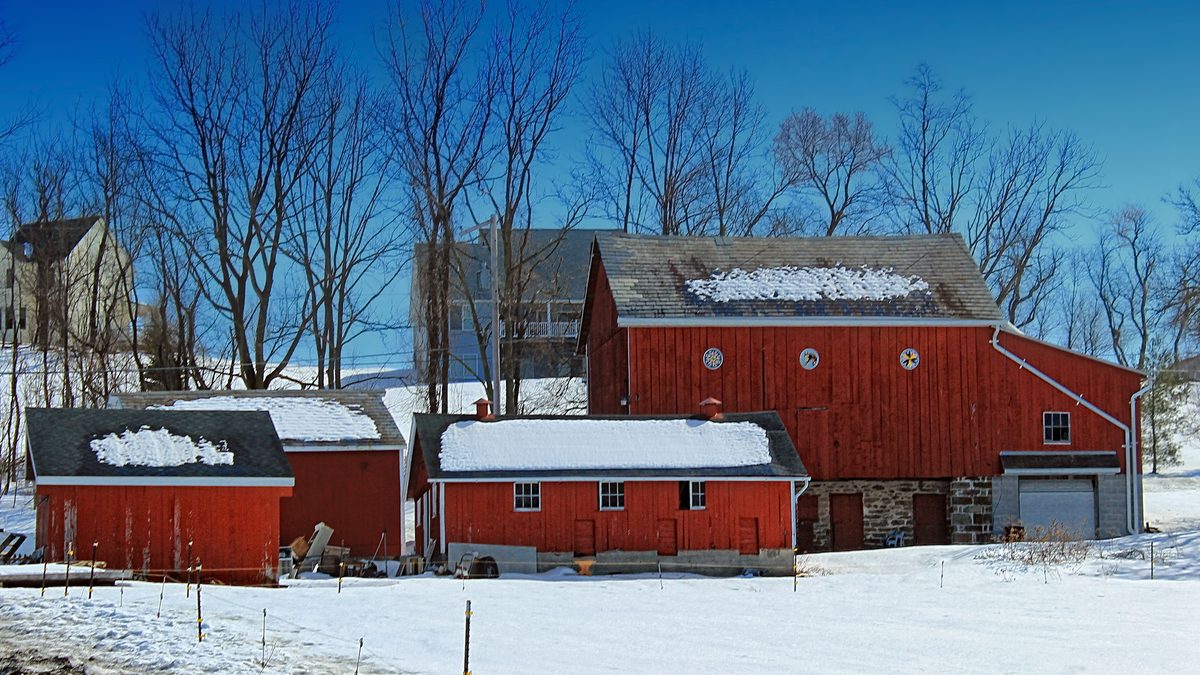 A red Pennsylvania bank barn and outbuildings under deep snow, with bare trees and a clear blue sky behind