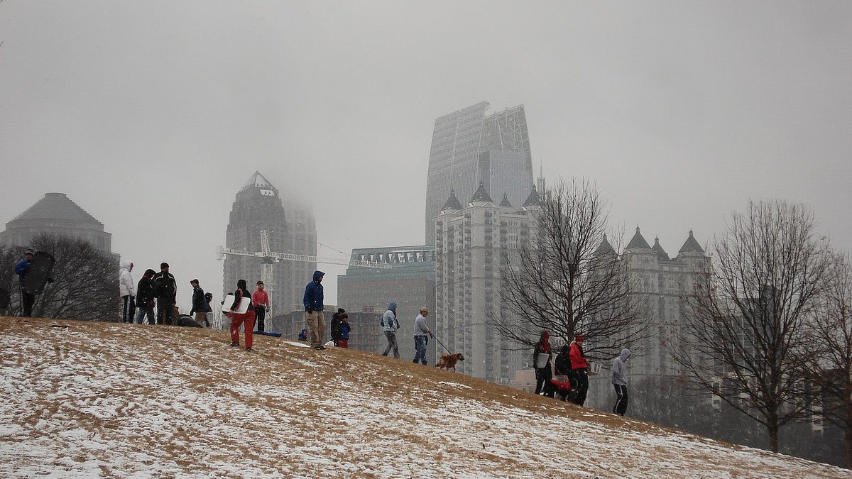 People trying to sled on a mostly bare grassy slope in Piedmont Park with the Atlanta skyline behind them during a light snowfall